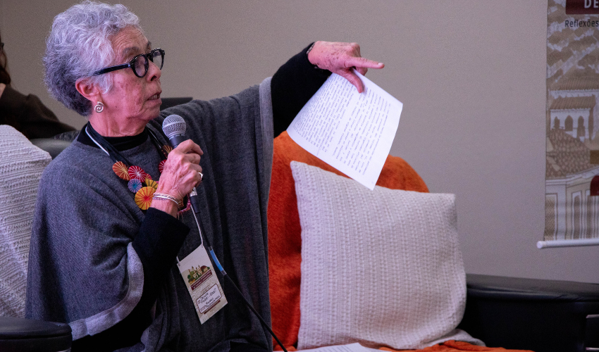 Idosa de óculos e cabelo grisalho fala ao microfone enquanto aponta, segurando folhas impressas, sentada em poltrona com mantas coloridas.
