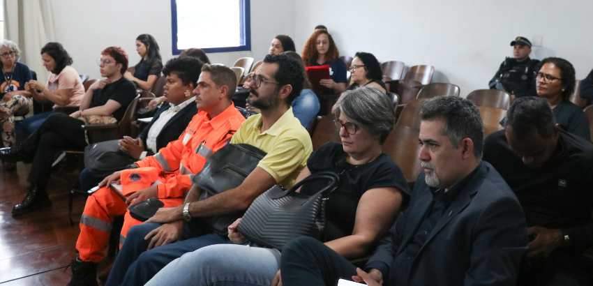 Pessoas sentadas em auditório durante um evento; à frente, profissionais de uniforme laranja e outros participantes atentos, incluindo mulheres e homens com bolsas e cadernos, enquanto policiais observam ao fundo