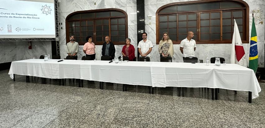 Foto de uma mesa de conferência com seis pessoas em pé atrás dela em um salão com decoração clássica, durante um evento educacional, com uma bandeira do Brasil e da instituição ao lado direito e uma apresentação projetada ao fundo com o título 'Curso de E