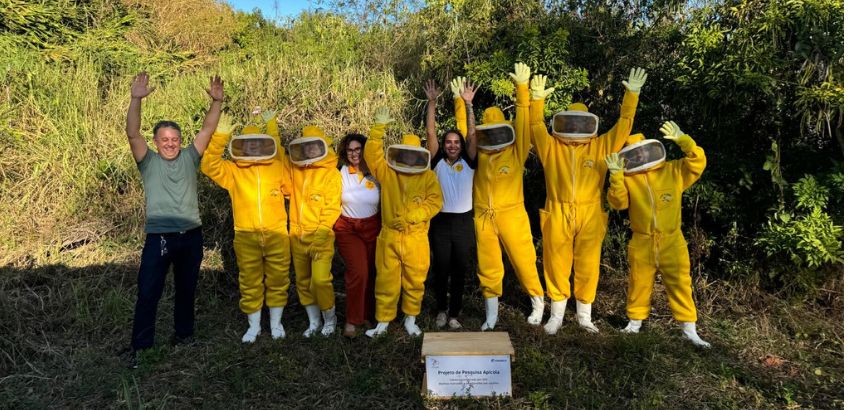 Foto de nove pessoas em um espaço aberto,seis com macacão amarelo e proteção de rosto e o restante com roupas comuns. Todos estão com os braços para o alto