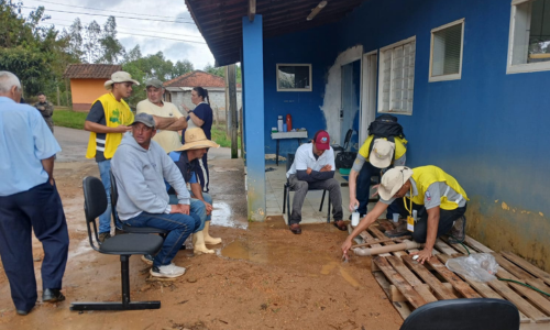 Foto de três estudantes de blusa amarela realizando uma oficina socioeducativa, dois deles ajoelhados demonstrando a atividade e um conversando, enquanto quatro moradores assistem