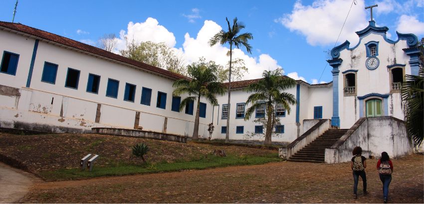 Fachada do prédio histórico do Instituto de Ciências Humanas e Sociais, com janelas e detalhes azuis, escadaria central, palmeiras ao redor e duas pessoas caminhando em direção à entrada sob céu azul com nuvens.