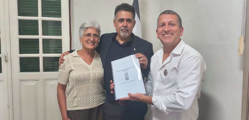 Três pessoas posam sorridentes em ambiente interno. A mulher à esquerda usa blusa clara; o homem ao centro veste blazer escuro; o homem à direita segura um documento encadernado entregue ao grupo. Ao fundo, portas brancas e parte de uma bandeira.
