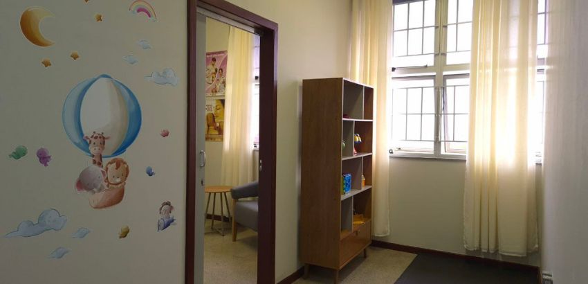 Sala iluminada com janelas amplas e cortinas claras, estante de madeira com brinquedos e, ao lado, outra sala com poltrona. Na parede externa, ilustração infantil mostra balão com animais e nuvens coloridas.