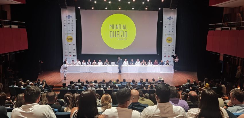 Auditório cheio durante o Mundial de Queijos do Brasil, com público sentado assistindo a uma mesa de jurados no palco. Ao fundo, um telão exibe o nome do evento. O ambiente é amplo, iluminado e formal, com pessoas acompanhando atentamente a apresentação.