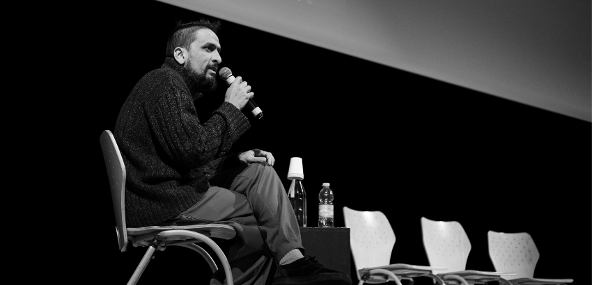 Homem sentado em uma cadeira no palco, segurando um microfone enquanto fala. À frente dele há uma mesa pequena com uma garrafa de água e um copo. Ao fundo, três cadeiras vazias alinhadas. Foto preto e branco.