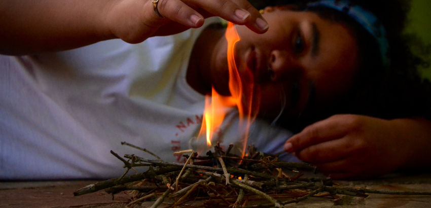 Menina deitada no chão observa uma pequena fogueira feita com gravetos, enquanto aproxima a mão do fogo.
