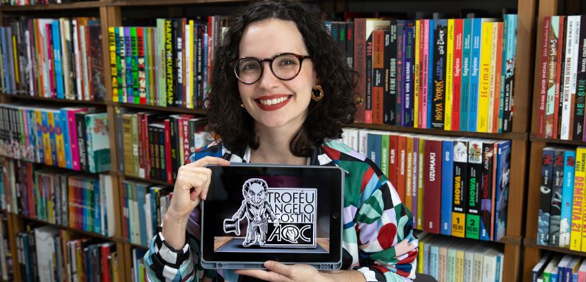 Mulher jovem de cabelos cacheados e óculos sorri enquanto segura uma placa com o logotipo do Troféu Angelo Agostini; ao fundo, estantes repletas de livros coloridos.