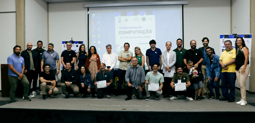 Grupo de participantes e organizadores posando para foto durante o IV Workshop da Computação, realizado nos dias 27 e 28 de novembro de 2019. As pessoas estão distribuídas na frente de um banner com o título do evento. Alguns participantes seguram certifi