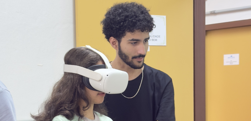 Duas pessoas em um ambiente interno; uma delas, uma jovem, usa um óculos de realidade virtual branco, enquanto a outra, um homem de cabelos cacheados, observa e parece orientá-la. O fundo tem paredes amarelas e brancas.