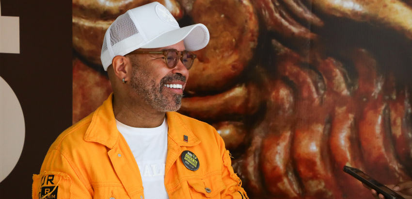 Dito pelo ChatGPT:  A imagem mostra um homem sorrindo enquanto usa uma jaqueta amarela, camiseta branca e boné branco. Ele está em um ambiente com uma parede de fundo que possui uma textura ou imagem abstrata. Ele também usa óculos e um bottom.