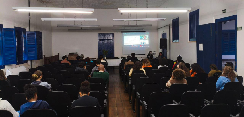 Imagem mostra um auditório com estudantes sentados e algumas cadeiras vazias. Os participantes do acompanham a apresentação via meet com professor assistente Manoel Horta Ribeiro, da Universidade de Princeton, EUA. O debate foi sobre mediação algorítimica