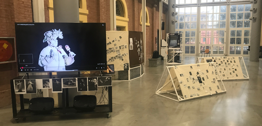 Instalação de videodança em um salão amplo, com telas exibindo vídeos em preto e branco, painéis inclinados e murais com fotografias. A sala tem grandes janelas ao fundo e iluminação natural