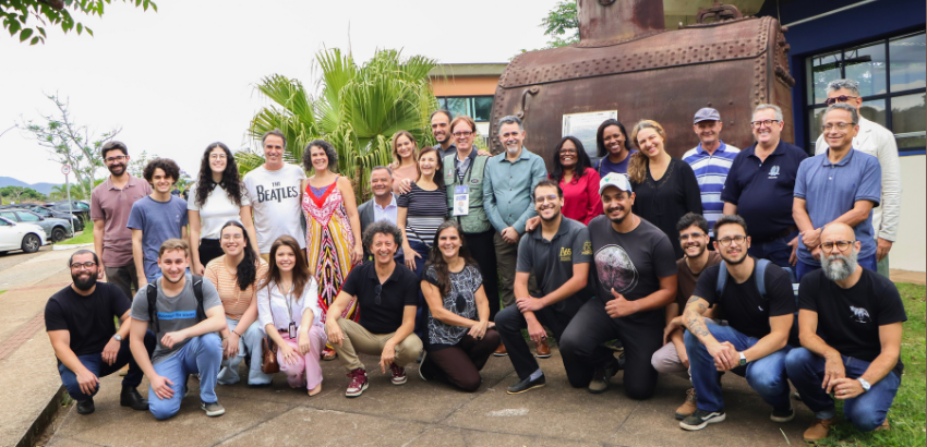 Grupo de pessoas posando para uma foto ao ar livre, em frente a uma estrutura metálica enferrujada e um edifício moderno.