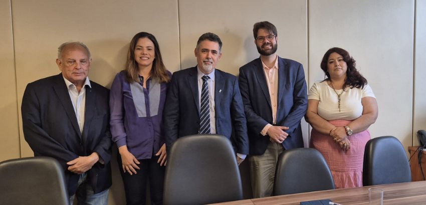Cinco pessoas posam lado a lado em uma sala de reuniões. Três homens e duas mulheres, todos vestidos com roupas formais. À frente deles, há uma mesa de madeira com cadeiras e alguns objetos, como um tablet, uma caneta e um copo.