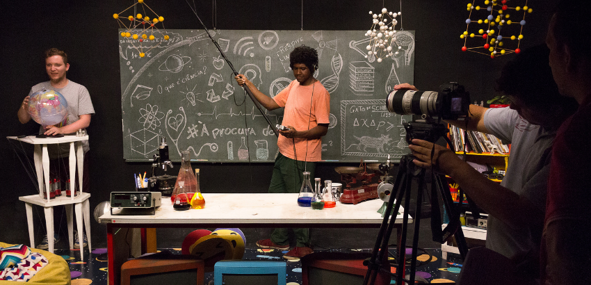 Cena em estúdio com mesa cheia de materiais de laboratório diante de um quadro com desenhos de ciência. Uma pessoa segura um globo e outra opera um boom. À direita, equipe filma com câmera profissional.