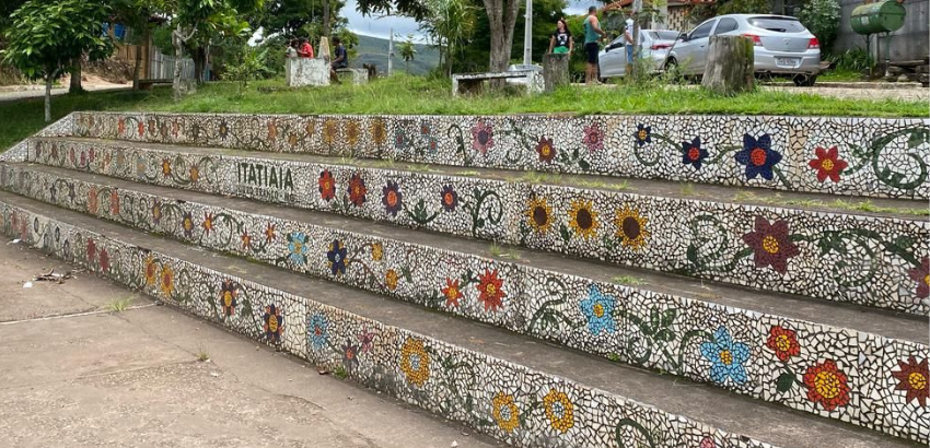 Escadaria ampla decorada com mosaicos de flores coloridas sobre fundo branco, localizada em área aberta com árvores ao redor. No degrau do meio, lê-se a palavra 'Itatiaia'. Ao fundo, há pessoas conversando, carros estacionados e vegetação verde.