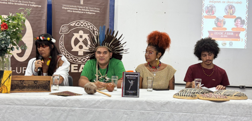 Imagem mostra a mesa do primeiro Adeando a UFOP.  Da esquerda para à direita: Raquel Pataxó, Cacique Danilo Borum-Kren, e os estudantes Brunely e Felipe da Pós- Graduação. Raquel e Danilo usam vestimetas tracionais indígenas. Eles estão numa mesa.