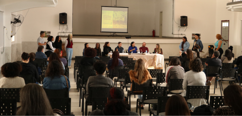 Em um auditório, público acompanha mesa de debate com representantes acadêmicos, lideranças comunitárias e movimentos. 
