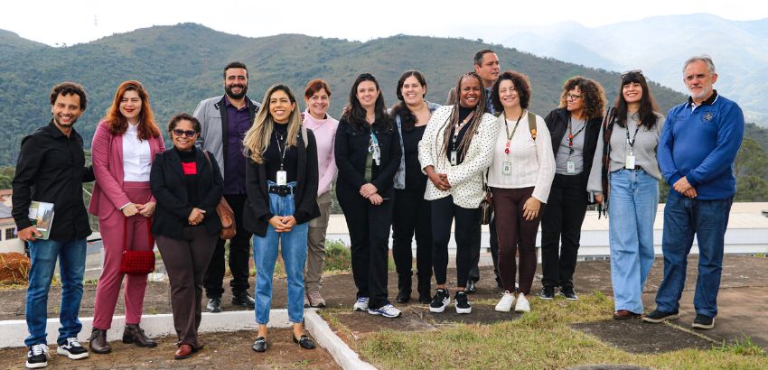 Grupo de 15 pessoas posa sorridente ao ar livre, com montanhas ao fundo. Elas estão lado a lado em pé, em área gramada, vestindo roupas casuais e crachás, indicando participação em visita técnica institucional.