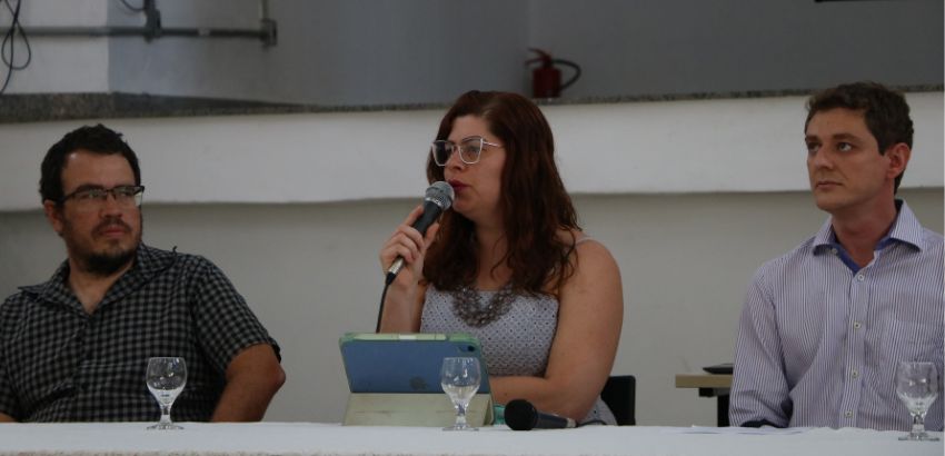 Três pessoas sentadas à mesa. A mulher no centro, de óculos, fala ao microfone. Dois homens, um de cada lado, a escutam.