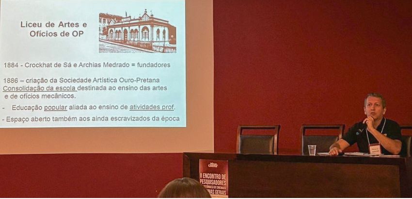 Homem fala em mesa de conferência durante apresentação de pesquisa. Ao fundo, projeção exibe slide sobre o “Liceu de Artes e Ofícios de OP”, com texto histórico e imagem antiga do prédio.