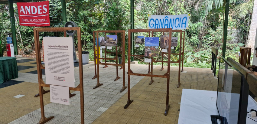 Visão geral da exposição Ganância, com painéis fotográficos, faixa com o nome da mostra e televisão exibindo vídeo em mesa lateral.
