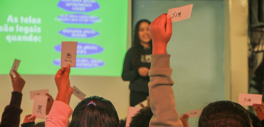 Alunos levantam cartões com a palavra "SIM" durante uma atividade interativa em sala de aula. Ao fundo, uma mulher apresenta conteúdo em um telão com a frase "As telas são legais quando:".