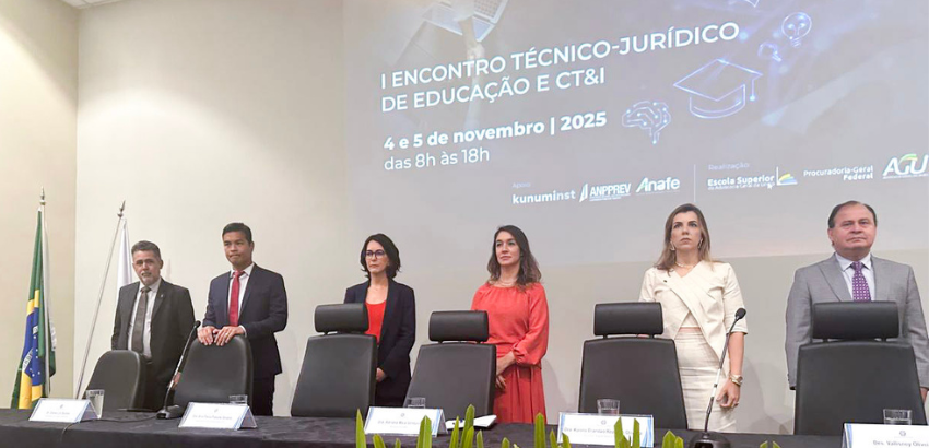 Pessoas em pé atrás de uma mesa no palco durante a abertura do “I Encontro Técnico-Jurídico de Educação e CT&I”, realizado em 4 e 5 de novembro de 2025, com flores à frente e o telão do evento ao fundo.