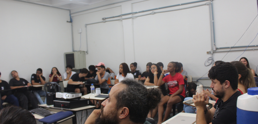 Sala de aula com estudantes ao fundo e palestrantes/professores em primeiro plano