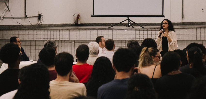 Professora ao fundo ministrando a aula com a plateia de costas assistindo à aula