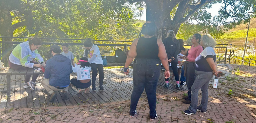 Grupo de pessoas reunidas ao ar livre sob a sombra de uma árvore, em atividade de observação e conversa, com paisagem rural ao fundo.