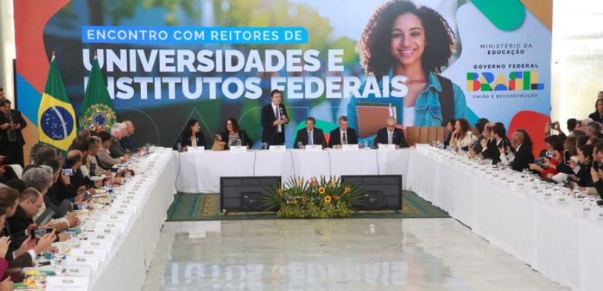 Encontro de reitores de universidades e institutos federais, com participantes sentados ao redor de uma mesa em formato de U, diante de um painel com o logotipo do Governo Federal e a imagem de uma jovem sorridente.