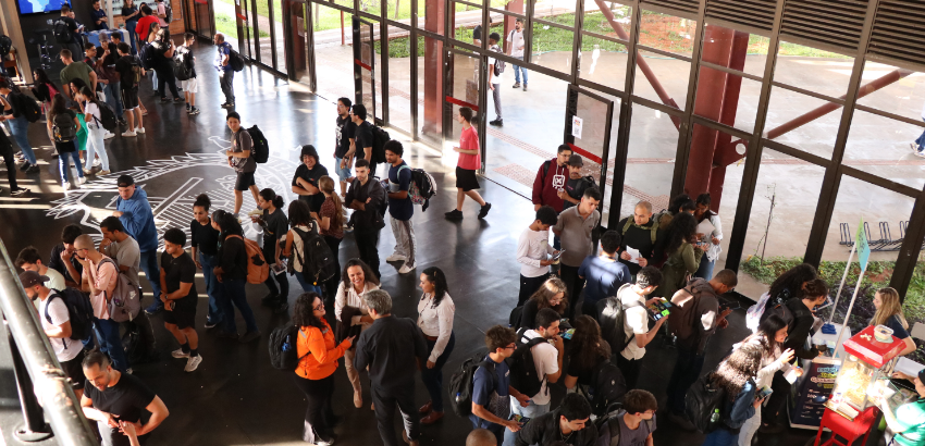 A imagem mostra um ambiente interno movimentado, com várias pessoas, muitas com mochilas, participando de um evento. Há filas, interações e um carrinho de pipoca, sugerindo um momento de recepção ou confraternização. O local tem piso escuro, paredes de vi