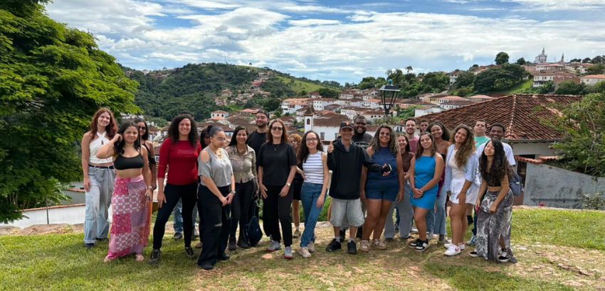 “Grupo de cerca de vinte jovens posando para uma foto em uma área gramada, com casas históricas de telhado vermelho e colinas verdes ao fundo, sob um céu parcialmente nublado.”