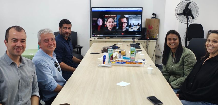 Seis pessoas participam de uma reunião híbrida em uma sala, quatro delas aparecendo em videoconferência na TV. Todos sorriem. Há lanches sobre a mesa. O ambiente é simples e iluminado.