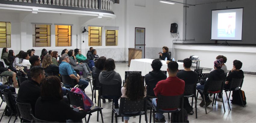 Auditório com cadeiras dispostas em semicírculo reúne estudantes e servidores durante palestra. Ao fundo, uma mulher fala ao microfone, sentada à mesa, com projeção na parede. Ambiente tem janelas com vidros coloridos e mezanino superior.