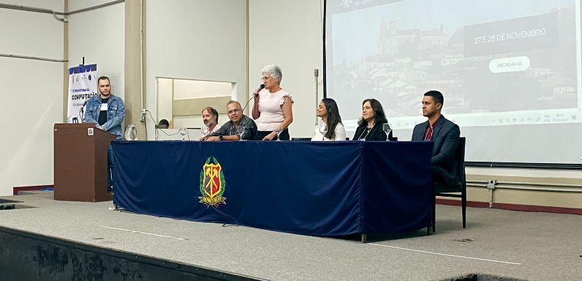 "Mesa de palestrantes no IV Workshop da Computação, com uma mulher de pé falando ao microfone e outros cinco membros da mesa sentados, incluindo homens e mulheres. Ao fundo, é possível ver um telão com a data do evento e um banner do workshop. Um dos pale