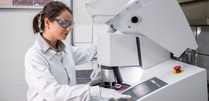 Pesquisadora em laboratório utiliza equipamento de análise metalográfica. Ela usa jaleco, luvas e óculos de proteção enquanto opera a máquina, demonstrando práticas seguras e precisas em ambiente científico e tecnológico