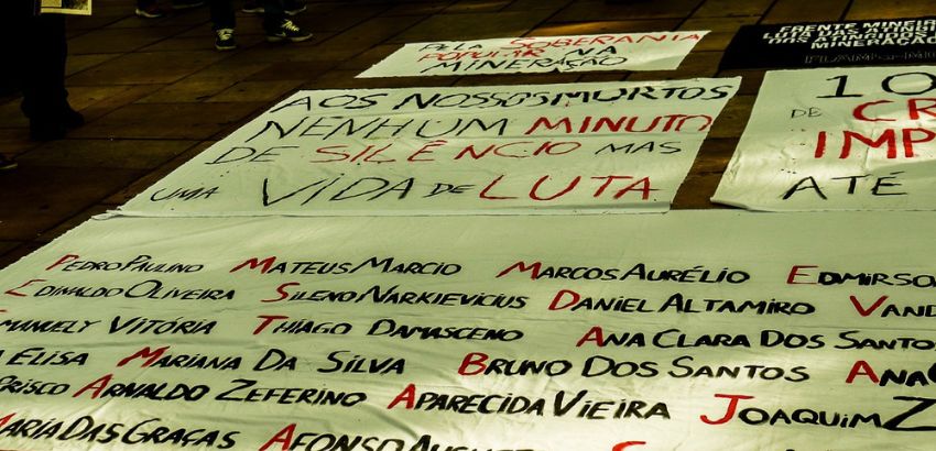 Faixas e cartazes estendidos no chão exibem mensagens de protesto e nomes de vítimas, com dizeres como “Aos nossos mortos, nenhum minuto de silêncio, mas uma vida de luta”. Manifestação denuncia crimes e pede justiça por tragédias ligadas à mineração.