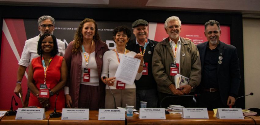 Sete pessoas estão em pé atrás de uma mesa de debate, sorrindo para a foto. Entre elas estão Joelma Gonzaga, Jandira Feghali, Laura Bezerra, Durval Ângelo, Luciano Campos e Cesar Callegari. Um banner vermelho compõe o fundo.