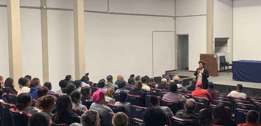 Auditório com público atento durante uma palestra; mulher em pé fala ao microfone diante de plateia diversa, sentada em cadeiras vermelhas e pretas. O ambiente tem paredes claras e iluminação artificial.        