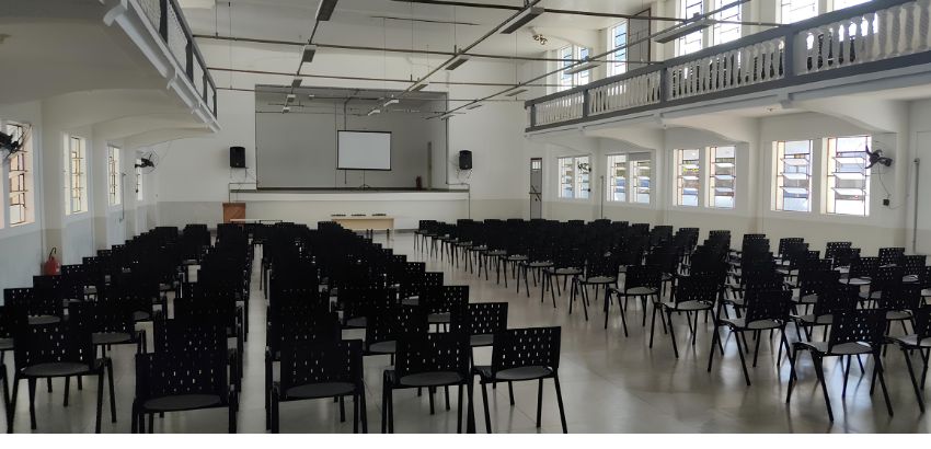 Auditório amplo e iluminado com fileiras de cadeiras pretas organizadas em blocos. Ao fundo, há um palco com projetor, tela branca, mesa e caixas de som. O espaço tem janelas grandes, piso claro e mezanino com guarda-corpo branco.