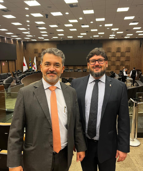 Foto de dois homens brancos centralizados usando terno, olhando para a frente e sorrindo, e ao fundo estando localizado o Tribunal Judicial de Minas Gerais
