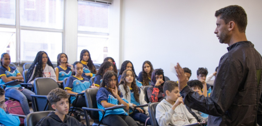 Imagem mostra o professor de matemática do Iceb Wenderson Marques Ferreira e os alunos da escola municipal sentados. Os alunos estão uniformes com na cor azul com detalhes em amarelho.