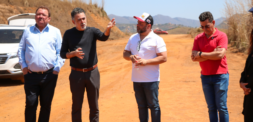 Um grupo de cinco homens conversa em uma estrada de terra em área rural sob céu claro. Um deles gesticula com a mão enquanto fala. Ao fundo, há carros estacionados e montanhas visíveis.