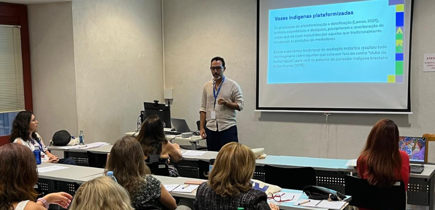 Homem apresenta palestra em sala de aula com projetor, enquanto várias pessoas sentadas assistem e fazem anotações. O slide no telão exibe o título "Vozes indígenas plataformizadas.