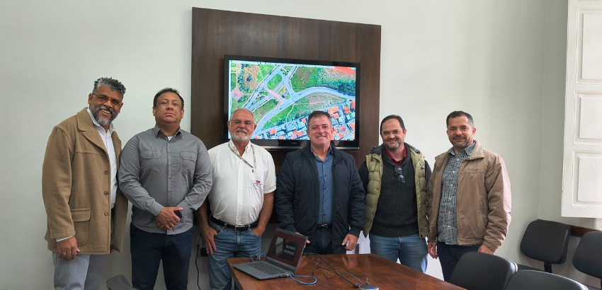 A imagem mostra seis homens em pé, lado a lado, posando para uma foto em uma sala de reunião. A sala é bem iluminada, com paredes brancas e uma janela grande à direita.