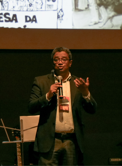 Homem falando ao microfone em um palco escuro durante uma apresentação. Ele veste blazer e camisa, gesticula com uma das mãos e aparece diante de um telão com imagem projetada ao fundo.