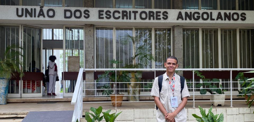 Imagem de Dalyson, estudante da UFOP em frente ao prédio da União dos Escritores Angolanos. O estudante está primeiro plano, com vestimentas em cores claras e um crachá. Observa-se ao fundo plantas e uma pessoa no sala de entrada.
